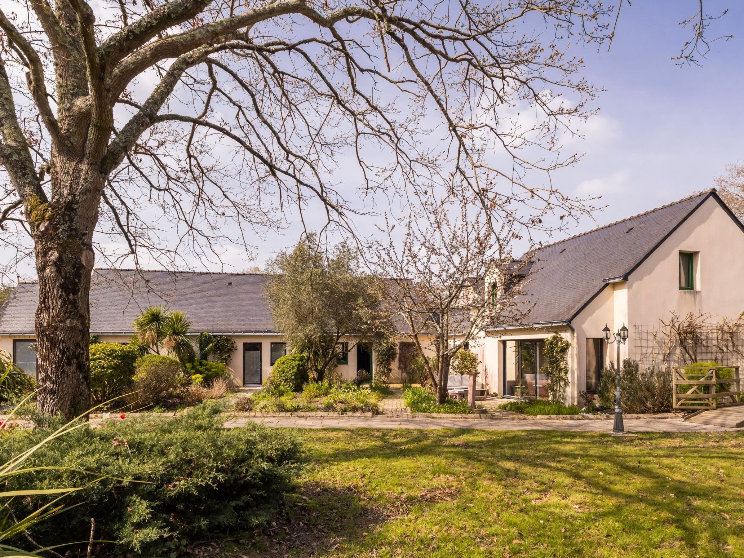 Een gebouw met twee verdiepingen met een schuin dak, een grote boom op de voorgrond en een verzorgde tuin met diverse struiken en gazon.