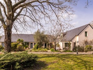 A two-story building with a sloped roof, a large tree in the foreground, and a well-maintained garden with various shrubs and a lawn. - Room with private terrace (Guérande)