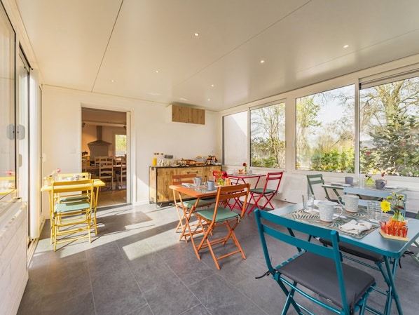 A modern dining area with a kitchen in the background, large windows, and a view of greenery.