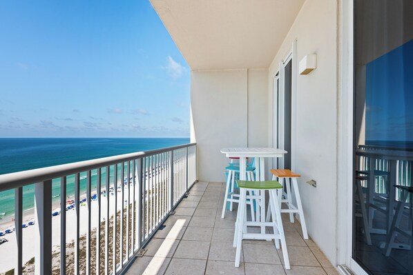 Un balcon avec vue sur la plage et lâocĂ©an, des tables et des chaises blanches, et une porte vitrĂ©e.