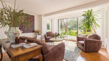 A modern living room with a wooden coffee table, two brown armchairs, a large vase with dried flowers, and a view of a garden through large sliding glass doors.