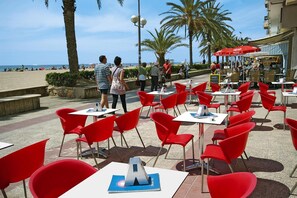 Terras met rode stoelen en tafels, een strand op de achtergrond en palmbomen.