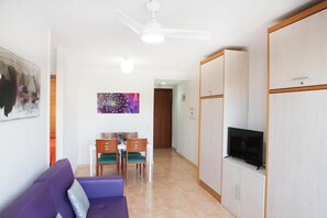 A living room with a purple sofa, a dining table with chairs, a ceiling fan, and a TV on a cabinet. - Apartamento en Calafell Cerca de la Playa (Calafell)