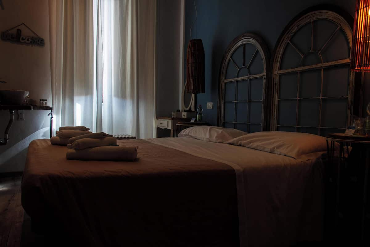 A dimly lit hotel room with a bed, a bedside table, a mirror, and a window with curtains.