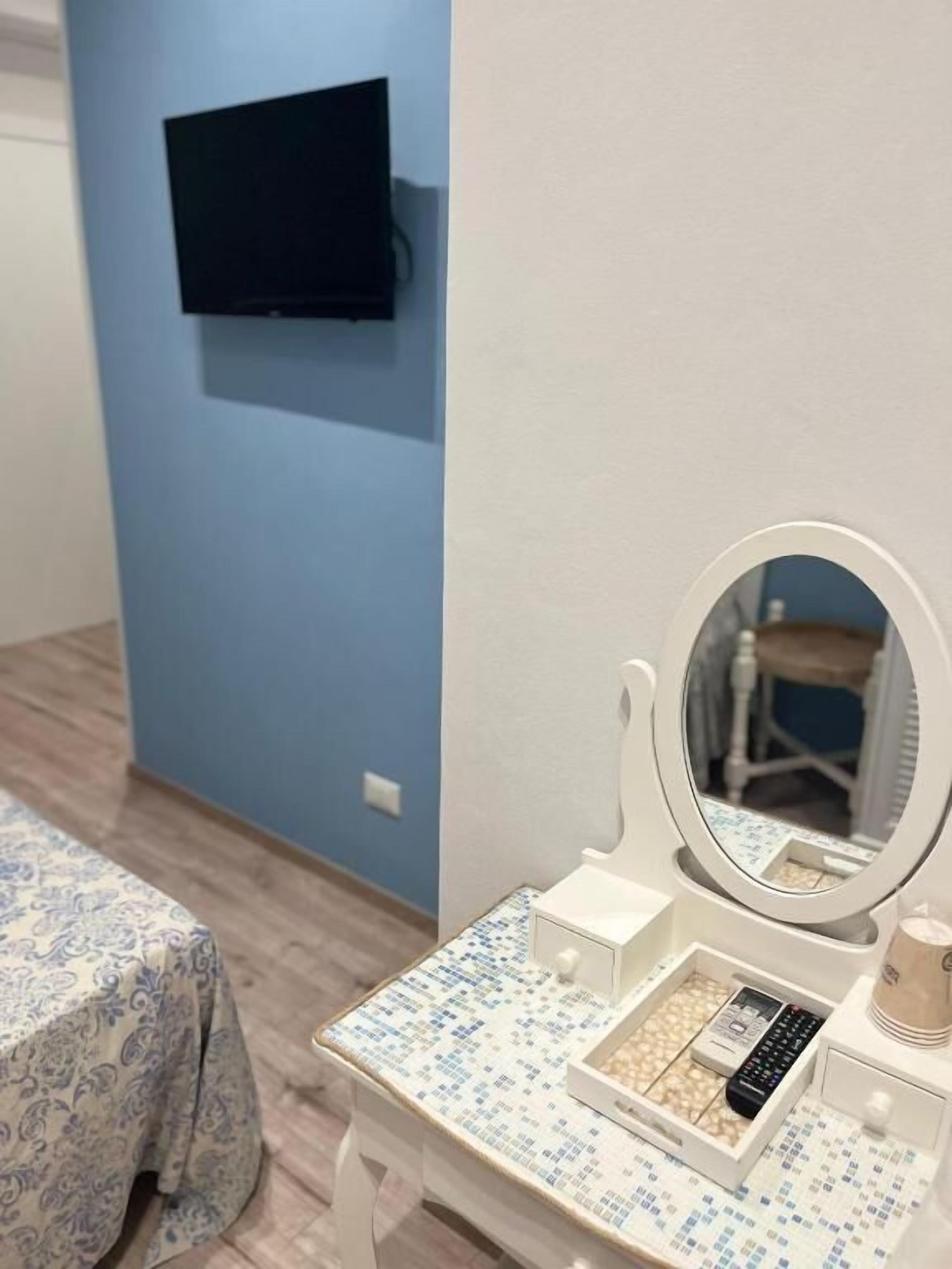 Ein Zimmer mit einem Bett, einem an der Wand befestigten Fernseher, einem Schminktisch mit Spiegel und einem kleinen Tisch mit Pflegeprodukten.