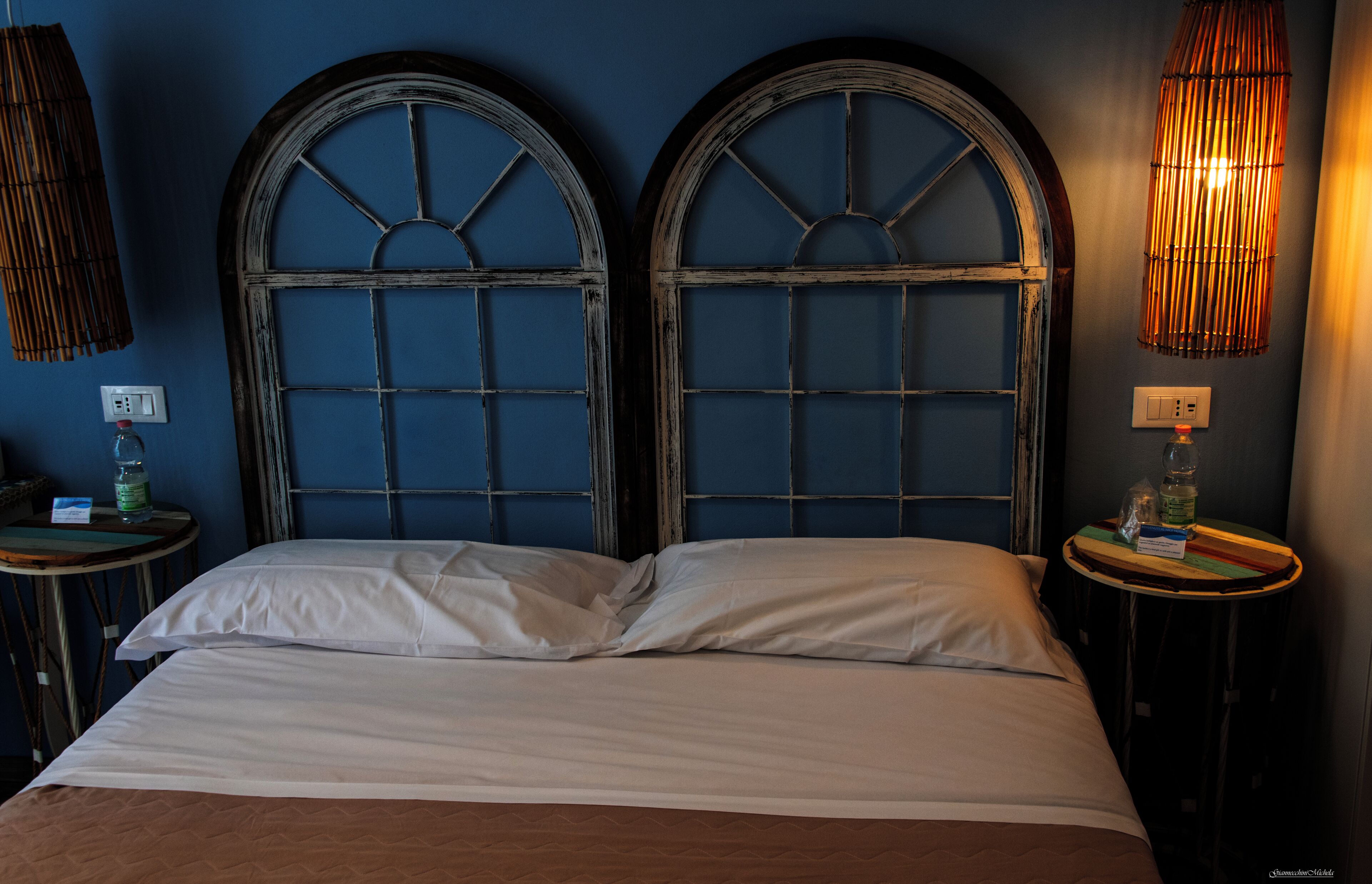 Ein Zimmer mit einem Bett, zwei Rundbogenfenstern, einer Wandlampe und einem kleinen Tisch mit einer Flasche und einem Glas.
