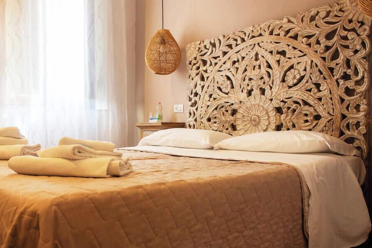 A neatly made bed with folded towels, a woven hanging light fixture, and a detailed carved headboard.