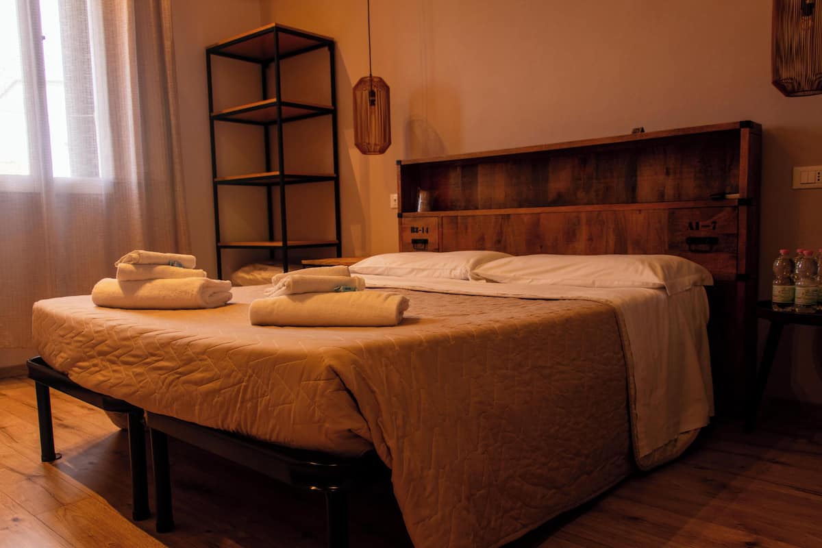 A neatly made bed with a wooden headboard, a bedside table with bottles, and a shelf unit against the wall.