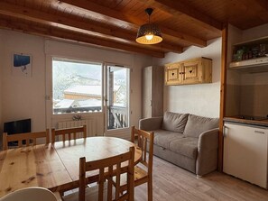 A cozy room with a wooden dining table and chairs, a sofa, a refrigerator, and a window with a view of snow-covered buildings. - Apartment mountain view - 4 guests (Crest-Voland)