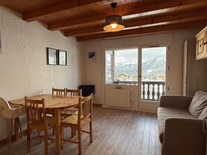A cozy room with a wooden dining table and chairs, a sofa, and a large window with a view of snow-covered mountains. - Apartment mountain view - 4 guests (Crest-Voland)