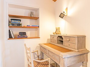 Ein Arbeitsraum mit Schreibtisch, Stuhl, Regalen voller Bücher, einem Teddybären und einer Wandleuchte.