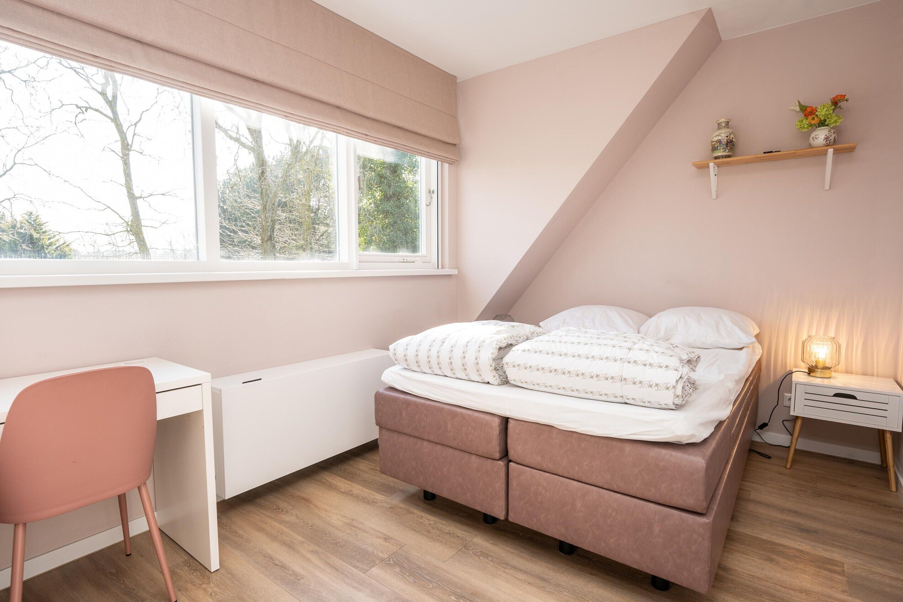 Une chambre à coucher comprenant un lit, un bureau, une chaise, un radiateur, une étagère avec des objets décoratifs et une fenêtre donnant sur des arbres.
