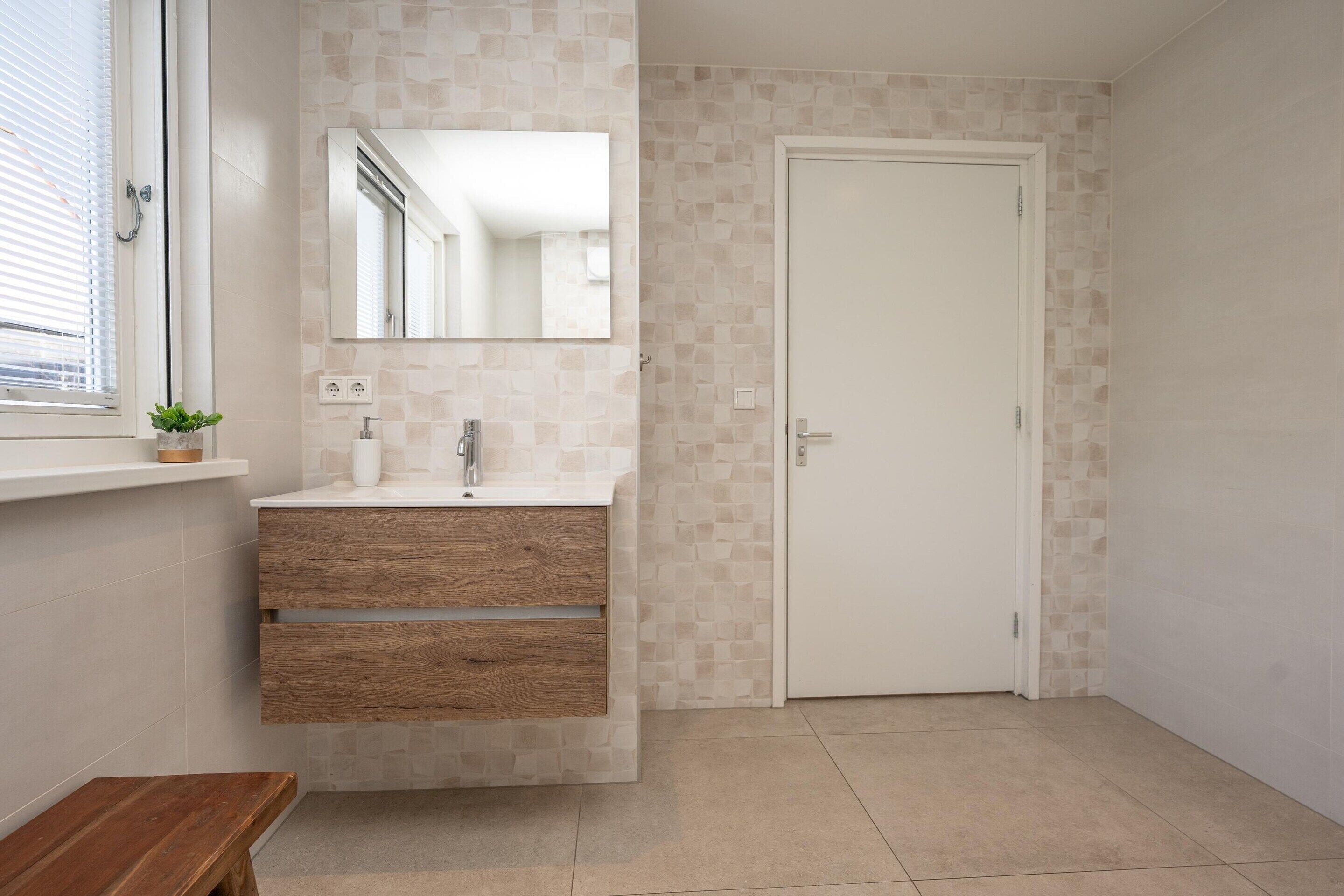 A bathroom with a wooden vanity, a mirror, a white door, and a small bench.