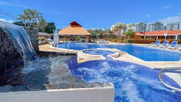 A pool area with a waterfall feature, lounge chairs, and a building with a red roof. - The Perfect Nest a Block Away to the Beach (Jacó)
