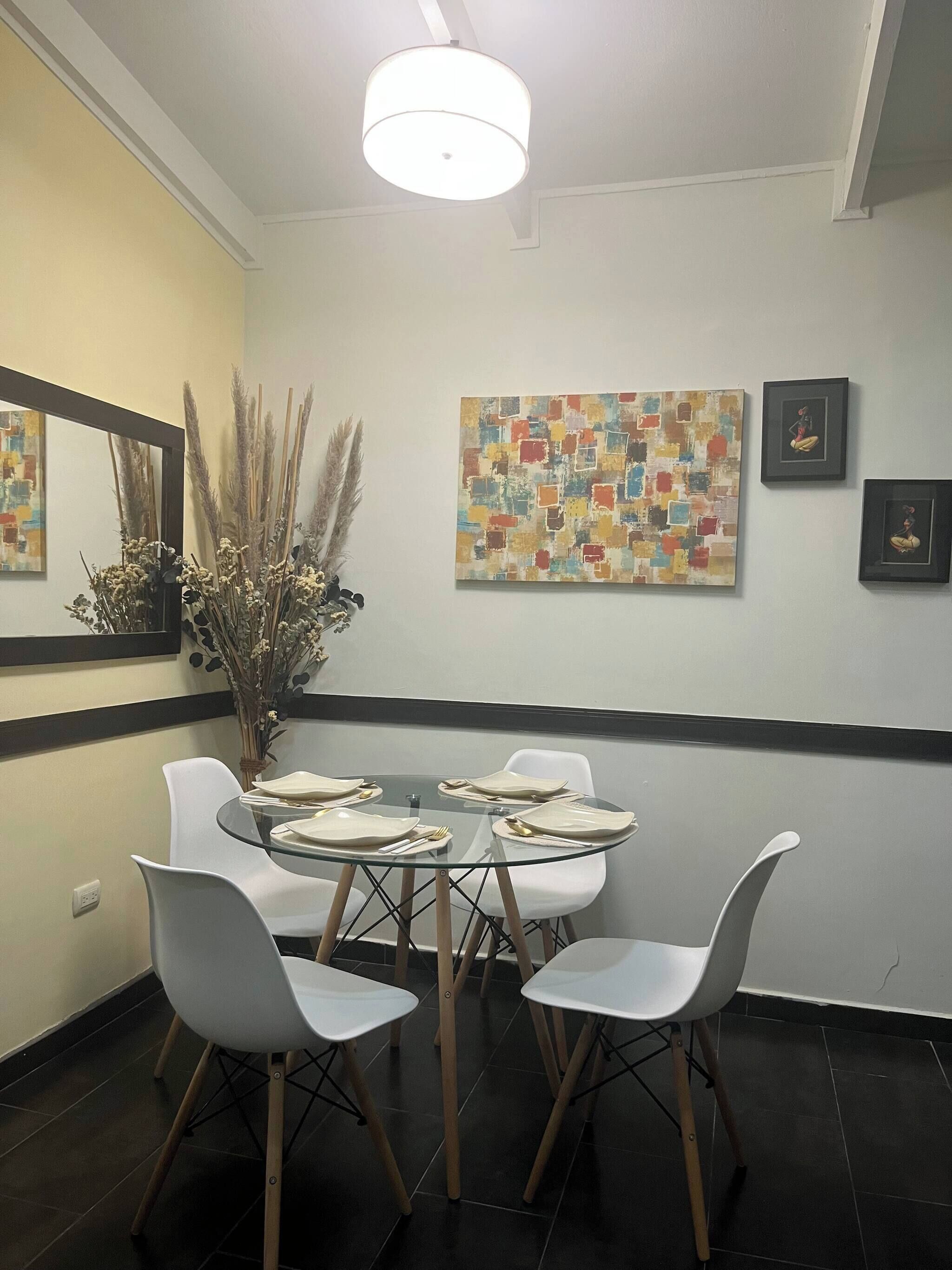 A dining area with a round glass table, white chairs, a vase with dried plants, and abstract wall art.