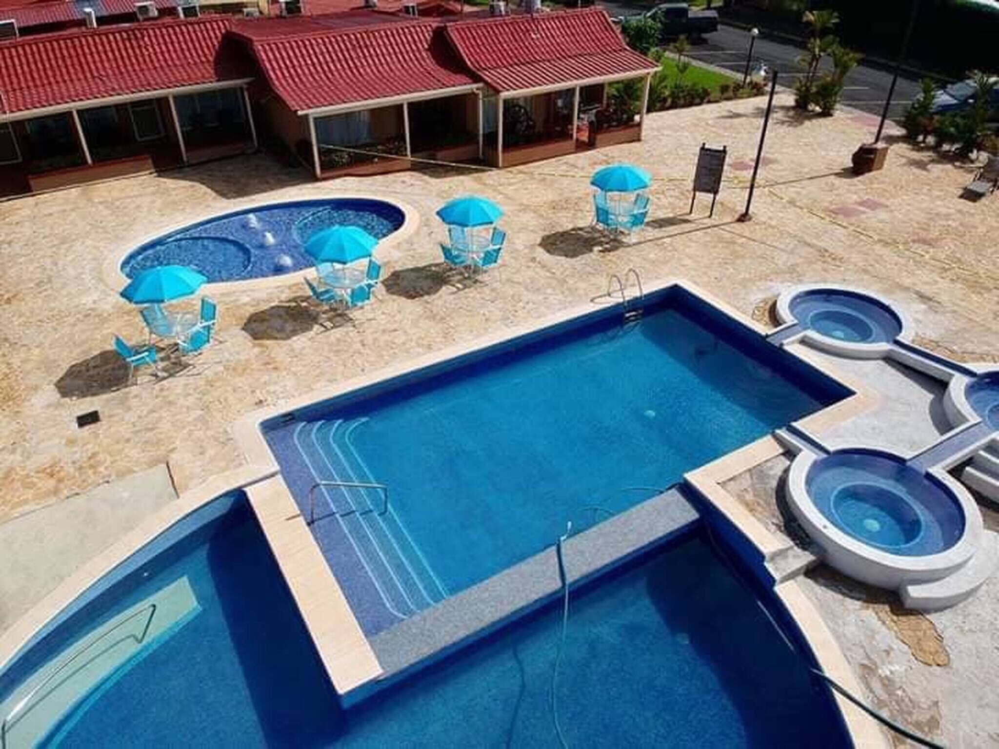 Uma piscina com várias áreas, incluindo uma piscina retangular principal e uma piscina circular menor, rodeada por espreguiçadeiras e guarda-sóis.