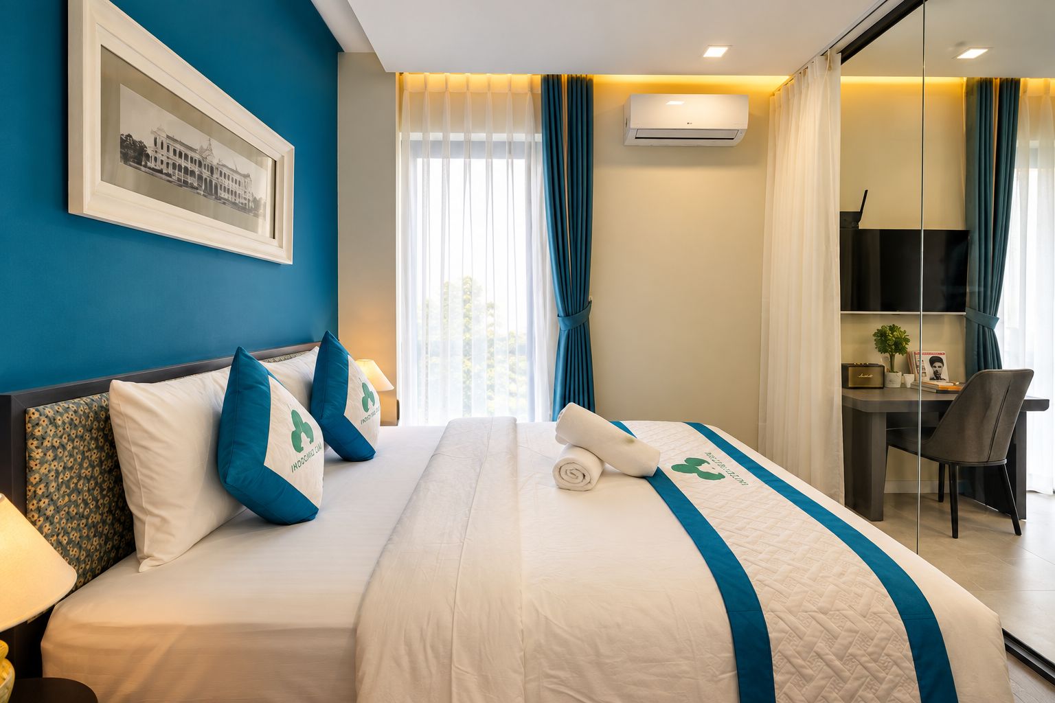 Ett hotellrum med en säng, ett skrivbord och en stol. Det finns ett fönster med gardiner, en luftkonditioneringsanläggning och en inramad tavla på väggen.