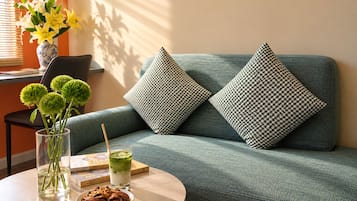 Una stanza con un divano verde, due cuscini decorati, un vaso trasparente con fiori verdi, un bicchiere di bevanda verde, un piattino con biscotti e un libro su un tavolo rotondo.