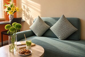 Ein Zimmer mit einem grünen Sofa, zwei gemusterten Kissen, einer klaren Vase mit grünen Blumen, einem Glas grünes Getränk, einer Schüssel Kekse und einem Buch auf einem runden Tisch.