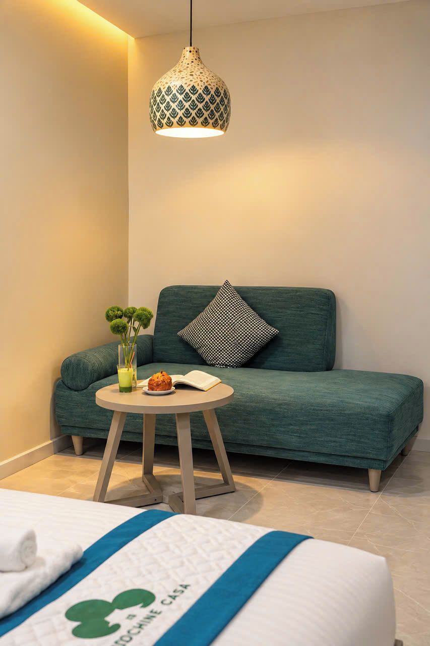 Una habitación de hotel moderna con un sofá verde, una mesita redonda pequeña con un vaso de jugo y un muffin, una lámpara colgante y una cama con una colcha azul y blanca.