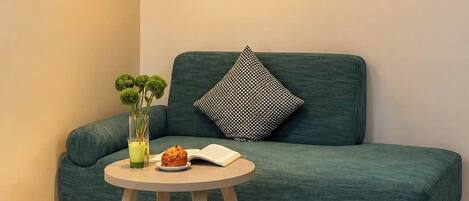 Una habitación de hotel moderna con un sofá verde, una mesita redonda pequeña con un vaso de jugo y un muffin, una lámpara colgante y una cama con una colcha azul y blanca.