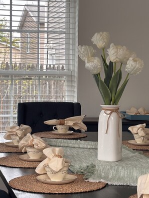 Ein Esstisch mit Platzsets, einer Vase mit weißen Blumen und einem Fenster mit Jalousien.