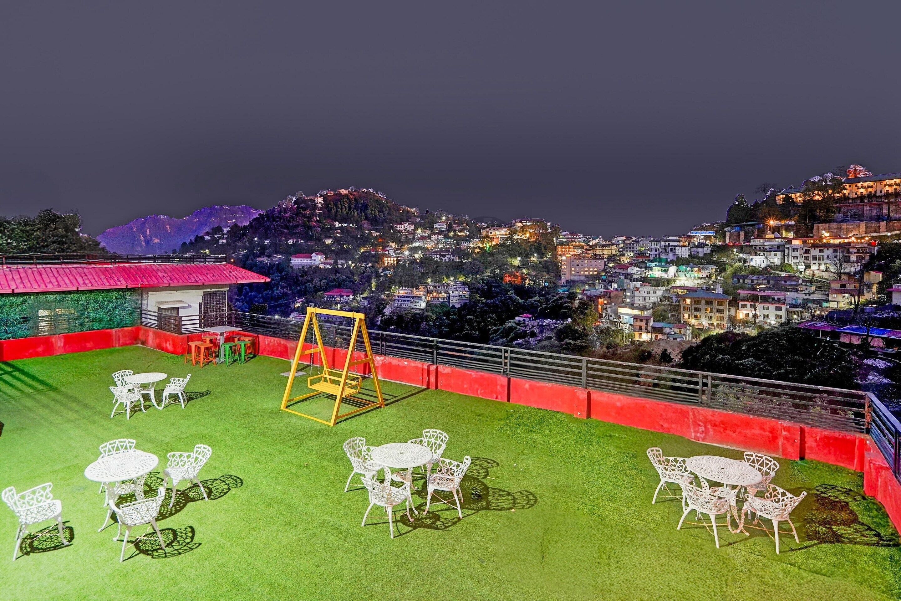 Una terraza en la azotea con mesas y sillas, un columpio y vistas de un paisaje urbano nocturno.