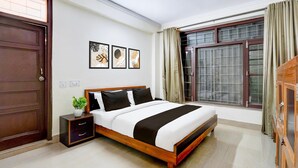 A neatly arranged bedroom with a wooden bed, a nightstand, a potted plant, and a window with curtains. - Townhouse Mall Road Near Library Chowk (Dehradun)