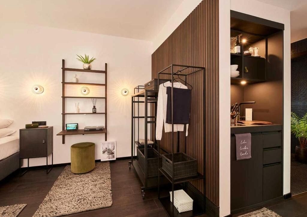 A modern room with a bunk bed, a wooden wall, a shelf with decorative items, a small round ottoman, and a sink area.