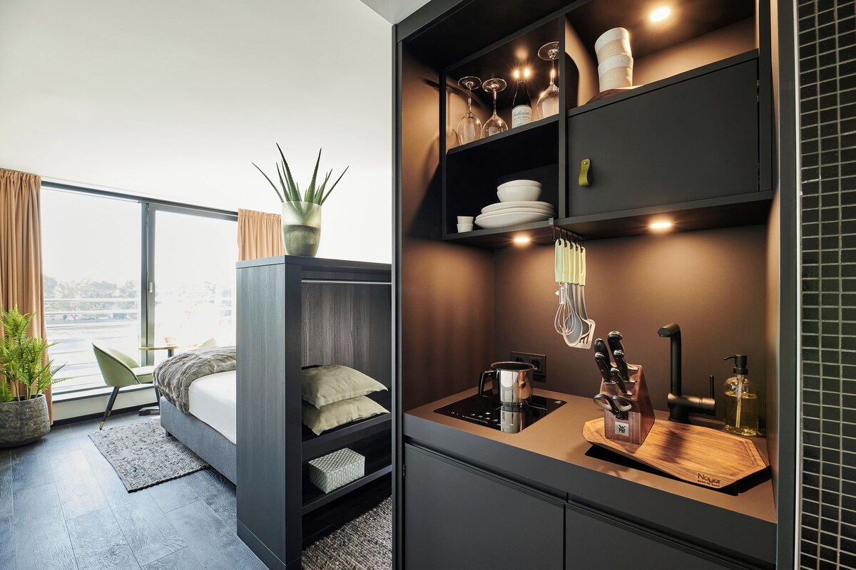 A modern kitchen with a built-in stove, a coffee machine, and a knife block. There is a plant on the counter and a view of the city through the window.
