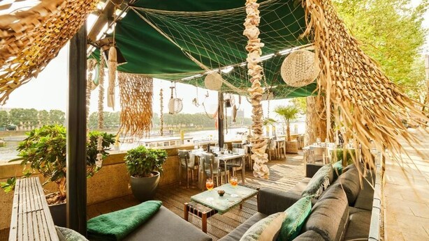 Un espacio al aire libre con una hamaca, mesas y sillas, rodeado de vegetación y vistas a la ciudad.