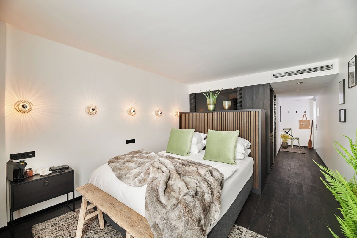 Un dormitorio moderno con una cama grande, mesitas de noche y un banco. Hay luces fijadas a la pared y una planta en maceta.