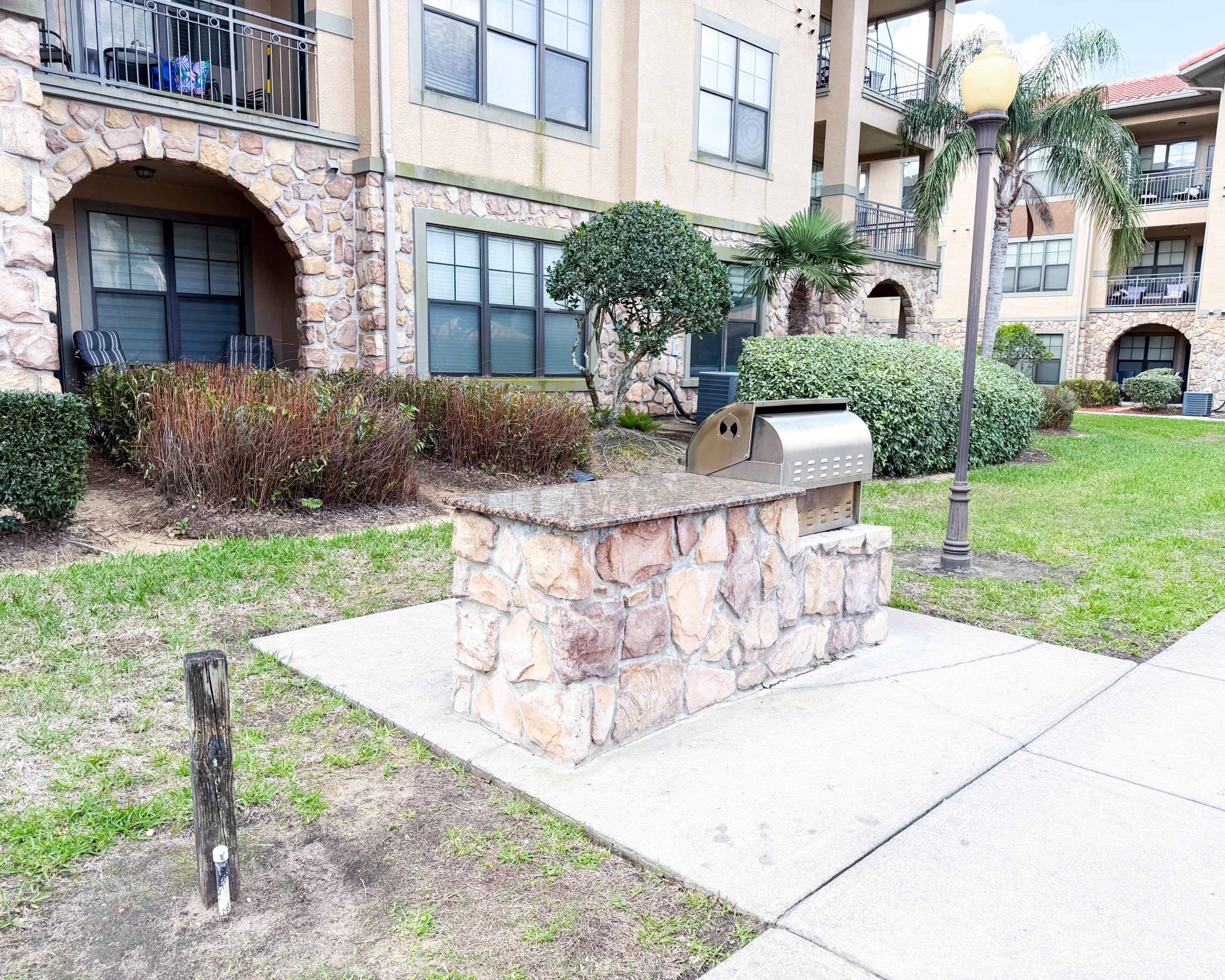 A stone-built outdoor grill area with a stone wall, surrounded by a well-maintained lawn and a multi-story building with balconies and windows.