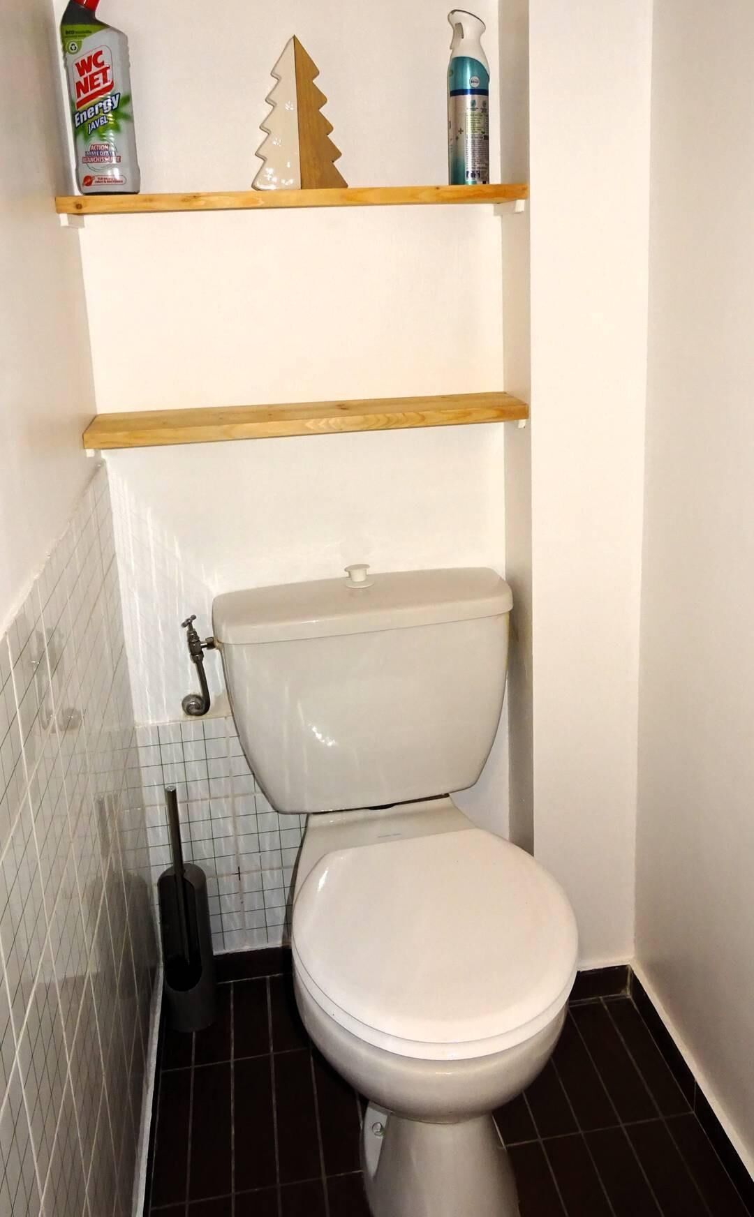 A toilet with a white seat and lid, a wooden shelf above it, and a white tiled wall.