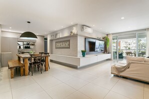 A modern living room with a sofa, a dining area with a table and chairs, a TV, and a large window. - 100m from beach | 3 suites | Barbecue area #CHA17 (Florianópolis)