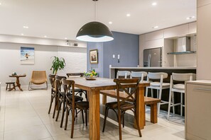 A modern kitchen with a dining area, featuring a wooden table, chairs, a hanging light fixture, and a refrigerator. - 100m from beach | 3 suites | Barbecue area #CHA17 (Florianópolis)