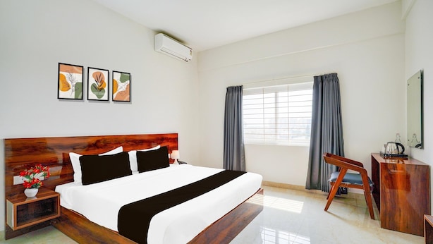 A modern hotel room with a large bed, a desk, a chair, a window with curtains, and artwork on the wall. - Deluxe Hebbal Townhouse (Bengaluru)