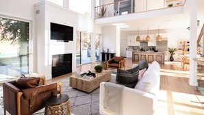 A spacious living area with a large TV, comfortable seating, a kitchen with modern appliances, and a balcony with bookshelves.