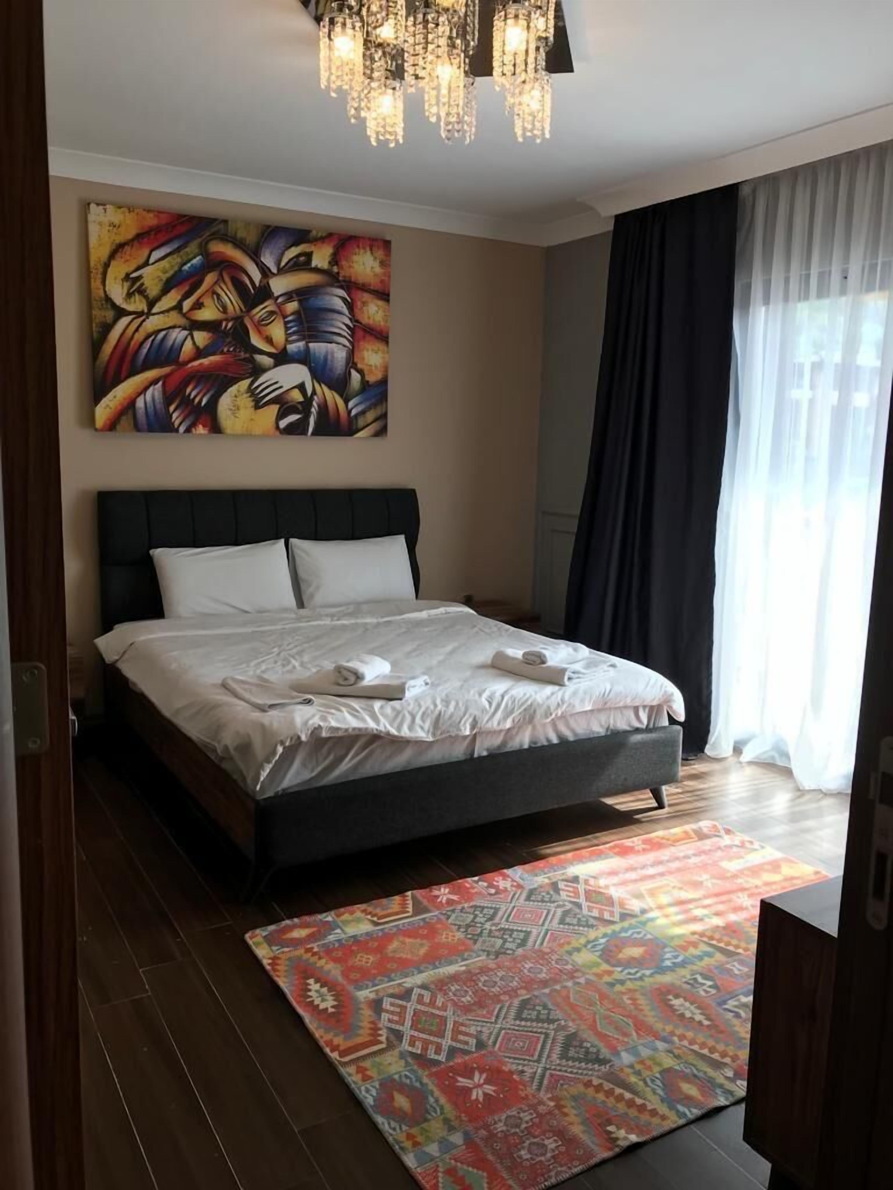 A modern hotel room with a large bed, a colorful abstract painting, a chandelier, and a patterned rug.