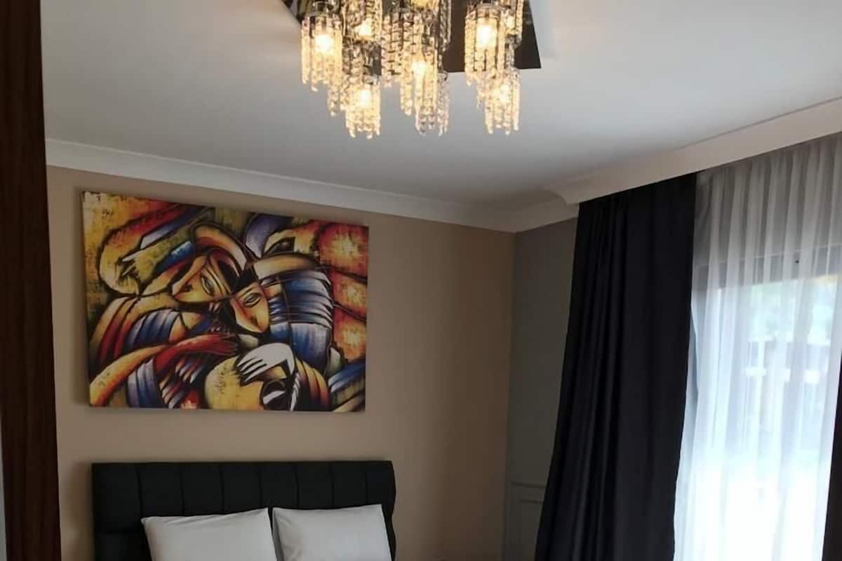 A modern hotel room with a large bed, a colorful abstract painting, a chandelier, and a patterned rug.