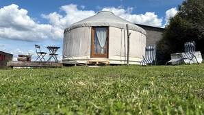 A yurt with a wooden door and a window, surrounded by a grassy field, with a building and chairs in the background. - Contemporary yurt (Hilsprich)