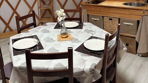 A dining area with a table set for four, a wooden bench, a hanging light fixture, a window with curtains, and a small shelf with a plant. - Contemporary yurt (Hilsprich)
