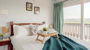 En pænt redt seng med en bærbar computer på et bakke bord, en sengetavlelampe, en plante og udsigt til det fri gennem en altan med gelænder.