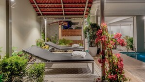 Et overdækket udendørs loungeområde med to liggestole, en pool og krukkeplanter.