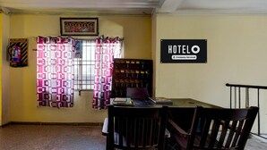 Quarto de hotel com uma mesa de jantar, duas cadeiras, uma janela com cortinas estampadas, uma placa na parede com a palavra 'HOTEL' e um extintor de incêndio na parede.