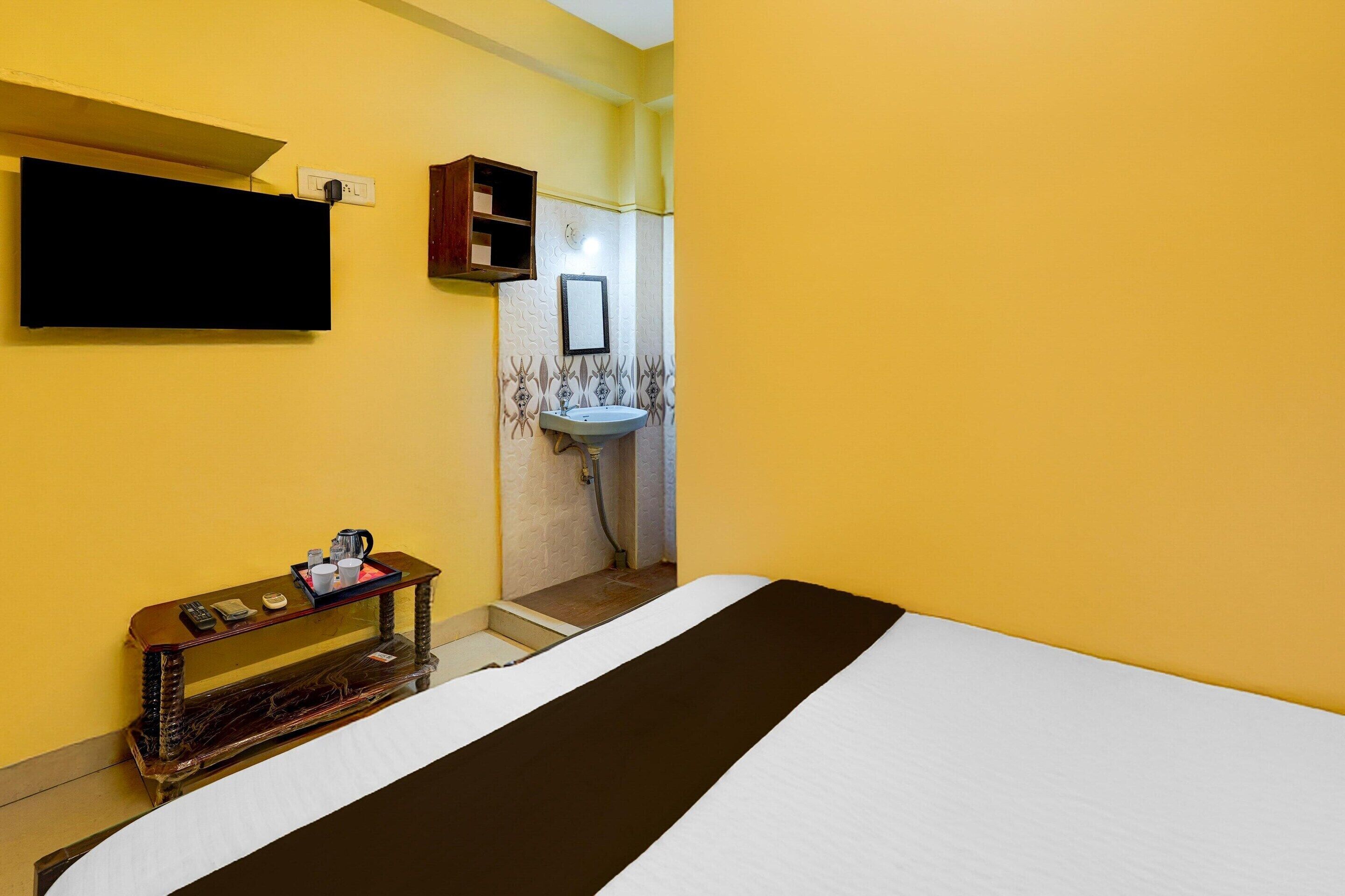A hotel room with a bed, a bedside table with a tray, a wall-mounted TV, a small shelf, and a bathroom with a sink and mirror.
