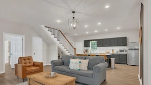 A modern living room with a sofa, coffee table, and a staircase leading to a kitchen area. - Hot Tub! Hidden Kayak Cove -2 Masters, Kayaks, Fire pit, & Game Room (Jasper)