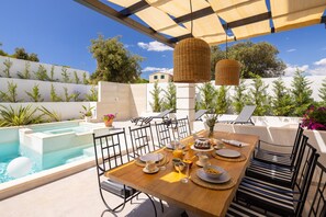 Uma área de refeições à beira da piscina, com mesa posta para o pequeno-almoço, piscina e uma pérgula com luzes pendentes.