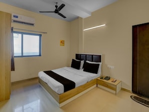 A bedroom with a bed, bedside table, nightstand, ceiling fan, window, and air conditioning unit. - Super Hotel O Vapi Gidc Formerly Zion Inn (Vapi)