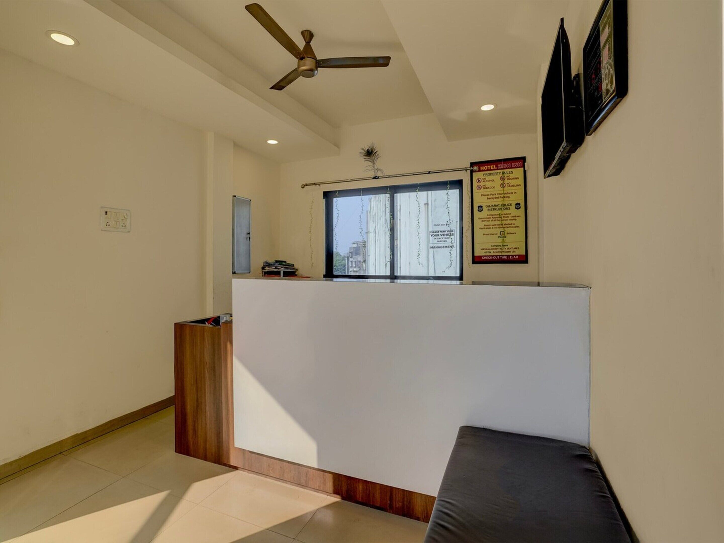 A hotel reception area with a reception desk, a ceiling fan, a window with a view of buildings, and a sign with property rules.